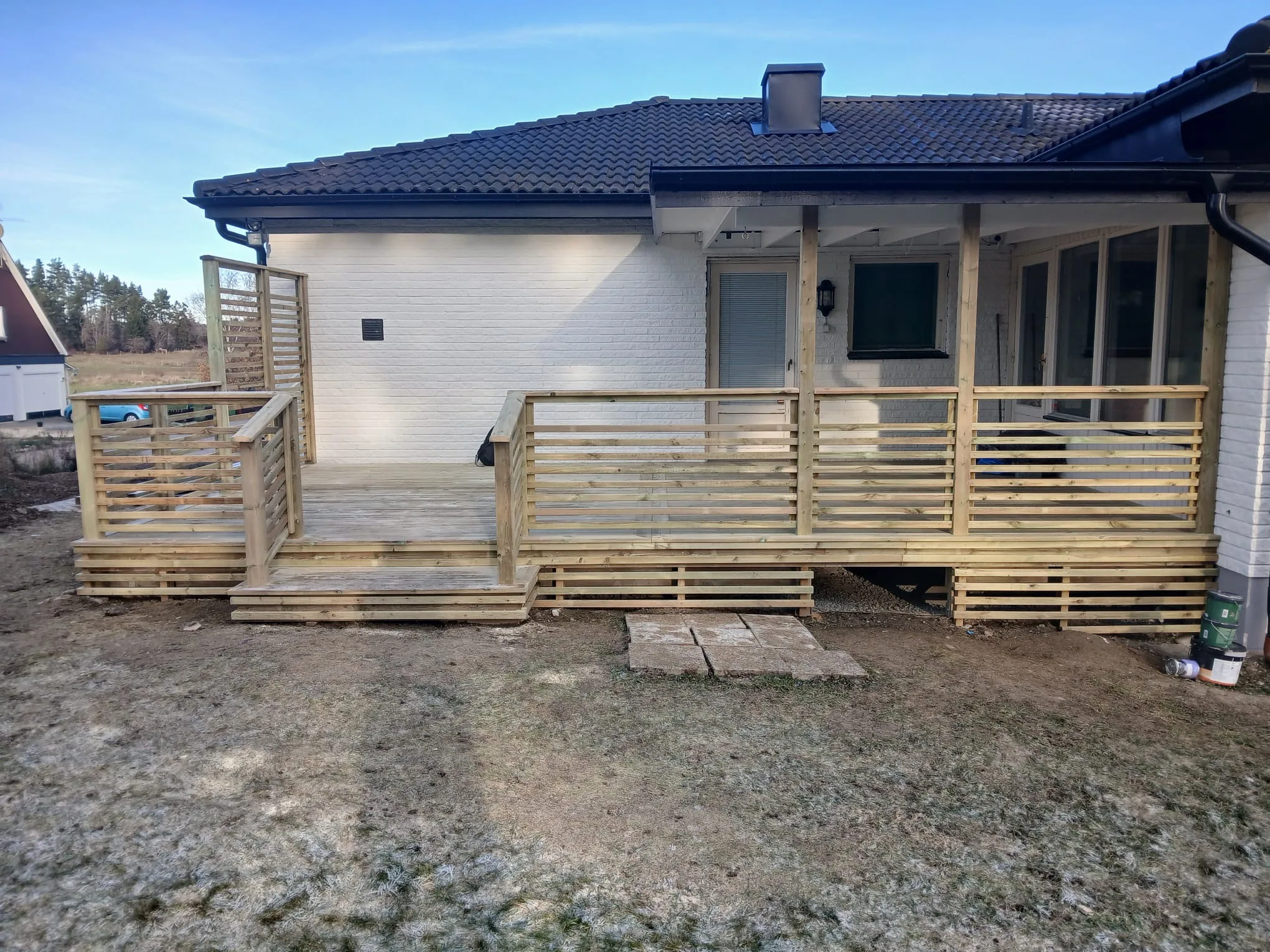 Trädeck med räcke och ramp vid entrén till huset, skyddat av omgivande landskap