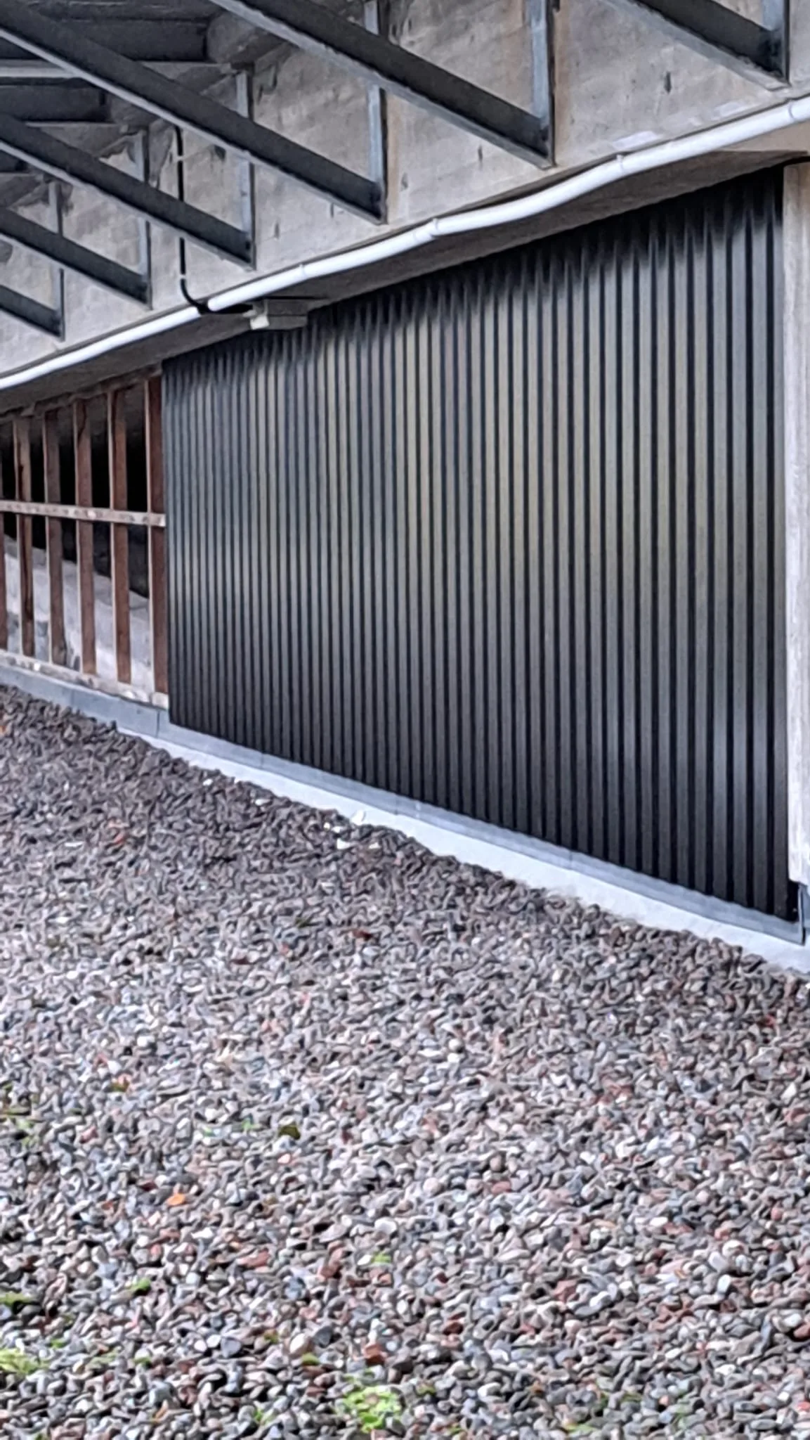 Svart vägg med vertikala ribbor under ett tak, med grusområde framför