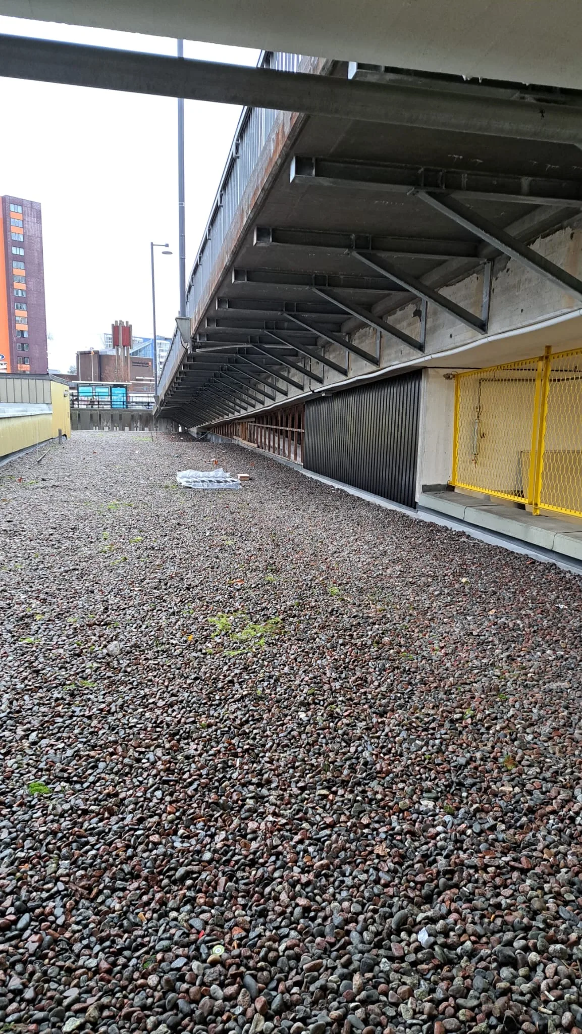 Grusområde under en byggnad med synliga konstruktionselement och säkerhetsstängsel